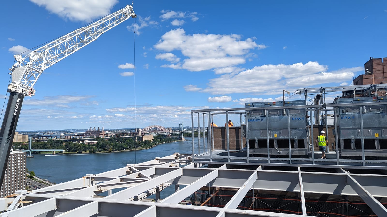Rooftop crane lift over structural steel with the East River and Hell Gate Bridge behind, New York
